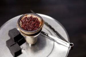 Close-up van een shishakop gevuld met rode waterpijptabak, met vier vierkante kooltjes en een metalen tang.