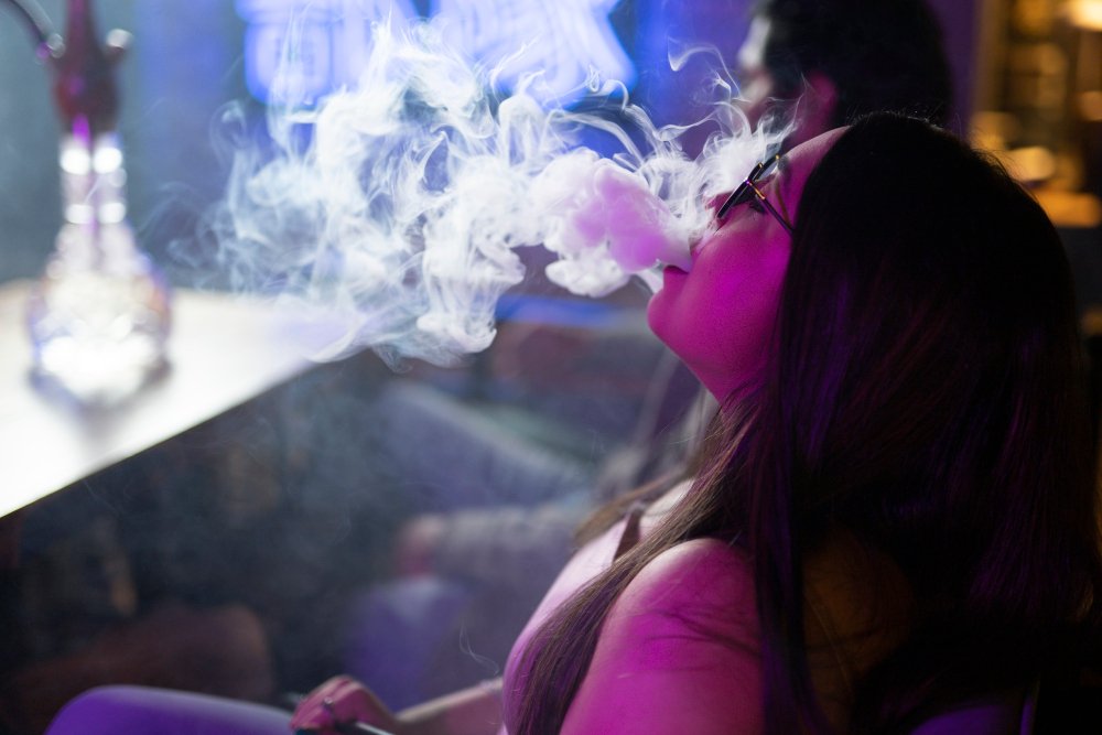 Vrouw blaast rook uit shisha (hookah) in sfeervolle, donkere bar.